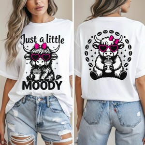Può includere: T-shirt bianco con grafica di una mucca delle Highlands con occhiali da sole a forma di cuore e fiocco rosa. Sul davanti c'è scritto "Just a little MOODY". Il retro presenta un design simile con chicchi di caffè.