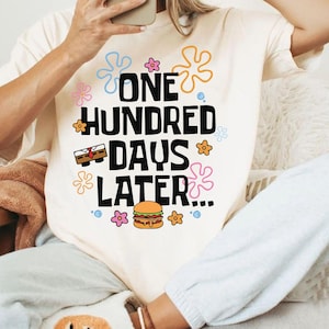 May include: Cream-colored t-shirt with the text "ONE HUNDRED DAYS LATER" in black, with cartoon graphics of a treasure chest, flowers, and a burger. The shirt is worn by a person.