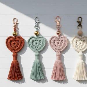May include: Four crochet heart keychains in shades of rust, mint, pink, and cream. Each heart has a matching tassel and a metal clasp. The keychains are adorned with a gold or copper-colored bead, and are displayed on a white wooden surface.