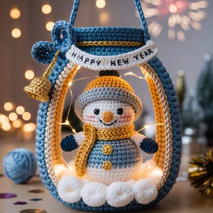 May include: A handmade crochet snowman inside a blue lantern with the words "Happy New Year". The snowman wears a yellow scarf and hat, with a blue and white body. The lantern has a gold bell and is lit with fairy lights.