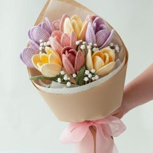 Op de afbeelding: Een boeket gehaakte tulpen in tinten geel, roze, paars en bordeaux, verpakt in bruin papier en vastgebonden met een roze lint. Het boeket bevat kleine witte vulbloemen.