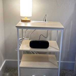 May include: A three-tiered, light wood and white metal side table with a drawer. A small lamp with a white shade and wooden base sits on the top shelf. A black speaker is on the middle shelf.