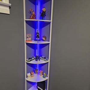 Puede incluir: Una estantería esquinera blanca con cinco estantes, iluminada por luces LED azules. Los estantes exhiben varias figuras, coches de juguete y objetos de colección. La unidad está situada en una esquina contra una pared gris.