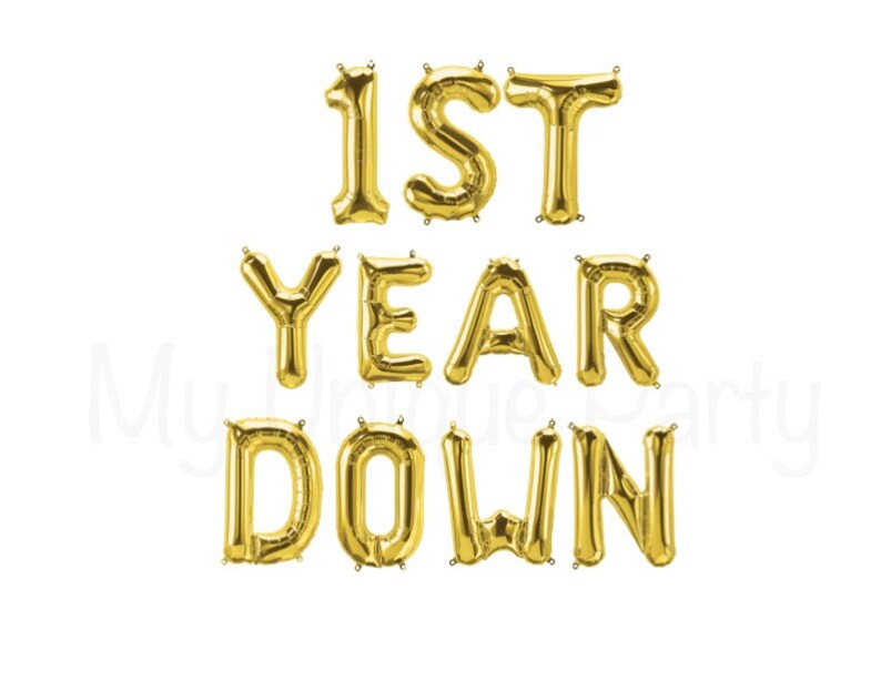 May include: Gold foil balloons spelling out "1ST YEAR DOWN" against a white background. The balloons are arranged to be read from left to right and top to bottom. Perfect for a first-year anniversary celebration.