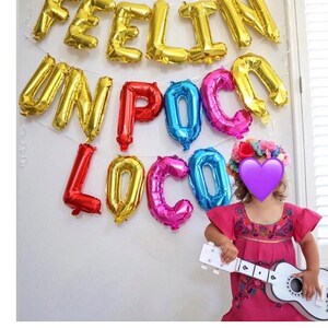 Pu&ograve; includere: Palloncini dorati formano la scritta "FEELIN' UN POCO LOCO" su un muro bianco. Sotto, una bambina con un vestito rosa e una corona di fiori tiene in mano una chitarra giocattolo bianca. I palloncini sono dorati, rossi, blu e rosa.