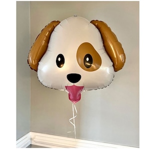 May include: A large, dog-shaped balloon with a white face, brown ears, and a brown spot on one side. The balloon has black eyes, a black nose, and a pink tongue. A pink balloon is attached to the bottom, with a white ribbon.