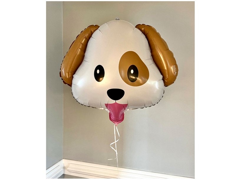 May include: A large, dog-shaped balloon with a white face, brown ears, and a brown spot on one side. The balloon has black eyes, a black nose, and a pink tongue. A pink balloon is attached to the bottom, with a white ribbon.