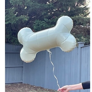 May include: A large, light green bone-shaped balloon floats in the air, held by a thin white string. The balloon is inflated and appears to be made of a shiny material. The background includes a wooden fence and green foliage.