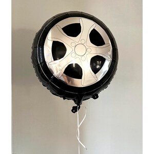 Pode incluir: Um bal&atilde;o preto em forma de pneu de carro com um design de roda prateada. O bal&atilde;o redondo est&aacute; inflado, com uma fita branca presa na parte inferior. O fundo &eacute; uma parede cinza clara lisa.