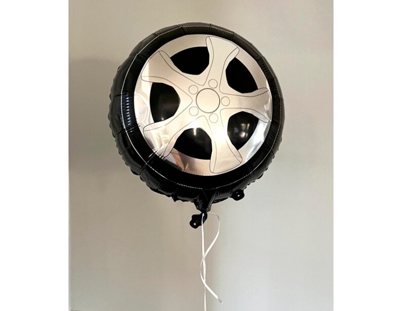 Pode incluir: Um bal&atilde;o preto em forma de pneu de carro com um design de roda prateada. O bal&atilde;o redondo est&aacute; inflado, com uma fita branca presa na parte inferior. O fundo &eacute; uma parede cinza clara lisa.