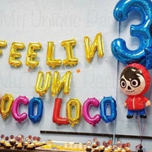 Pu&ograve; includere: Decorazione per festa di compleanno con palloncini in lamina che compongono la scritta "FEELIN' UN POCO LOCO". Un grande palloncino blu con il numero "3", un palloncino a forma di personaggio Coco e altri palloncini colorati. Cupcake sul tavolo.