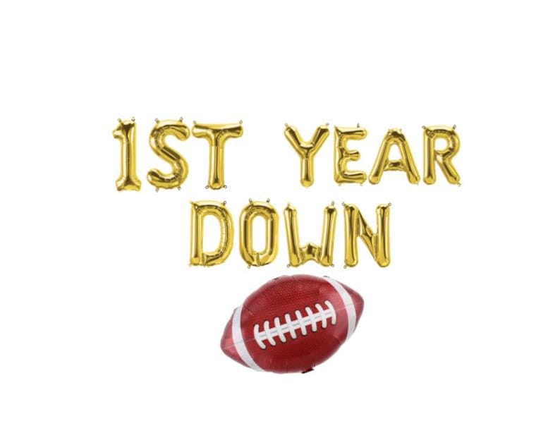 May include: Gold foil balloon letters spell "1ST YEAR DOWN" above a red and white football-shaped balloon. The balloons are set against a white background, creating a festive and celebratory theme.