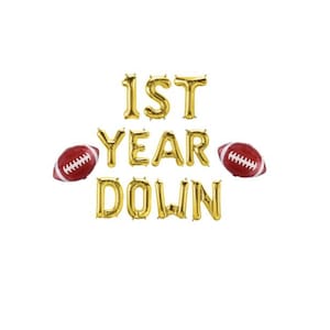 May include: Gold foil balloon letters spell out "1ST YEAR DOWN" with two red and white football balloons on a white background. A festive decoration for a first birthday or sports-themed celebration.