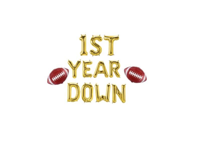 May include: Gold foil balloon letters spell out "1ST YEAR DOWN" with two red and white football balloons on a white background. A festive decoration for a first birthday or sports-themed celebration.