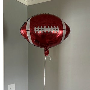 May include: A red and white football-shaped balloon. The balloon has white stripes and stitching details, and is suspended by a white string. The balloon is a festive decoration, perfect for a sports-themed party or event.