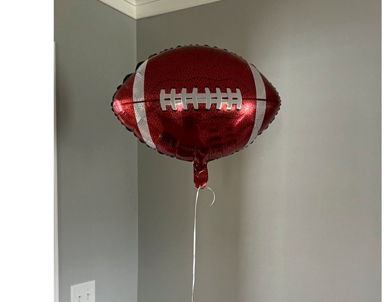 May include: A red and white football-shaped balloon. The balloon has white stripes and stitching details, and is suspended by a white string. The balloon is a festive decoration, perfect for a sports-themed party or event.