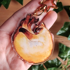 Può includere: Un portachiavi in ​​acrilico trasparente con un'illustrazione ad acquerello di un gatto siamese rannicchiato. Il portachiavi ha un ciondolo dorato a forma di stella attaccato a un anello diviso dorato.