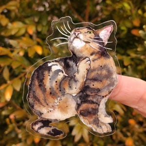 Peut inclure: Un autocollant transparent avec une illustration aquarelle d'un chat tigr&eacute; regardant vers le haut. Le chat est brun, noir et blanc avec un nez brun clair et des yeux verts.