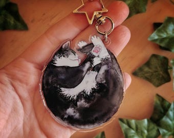 Puede incluir: Un llavero de acrílico transparente con un gato blanco y negro enroscado y durmiendo, pintado en acuarela. El llavero está unido a un amuleto de estrella plateado.