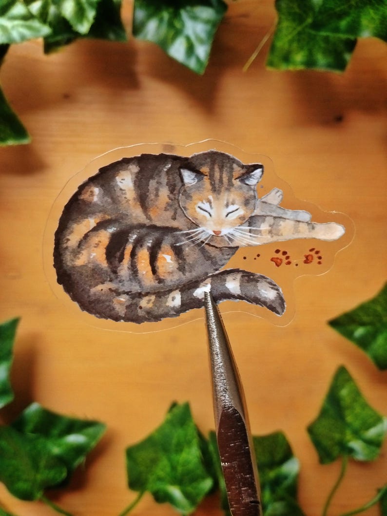 Peut inclure: Un autocollant transparent illustrant une aquarelle d'un chat tabby endormi avec des marques orange, noires et blanches. Le chat est enroul&eacute; en boule avec ses pattes rentr&eacute;es sous son corps. L'autocollant a une bordure blanche et est imprim&eacute; sur un fond transparent.