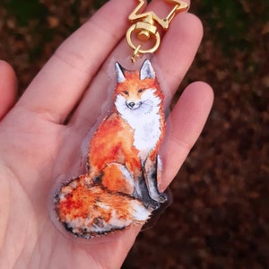 May include: A clear acrylic keychain featuring a hand-painted illustration of a red fox sitting with its tail curled around its body. The keychain has a gold star-shaped clasp.