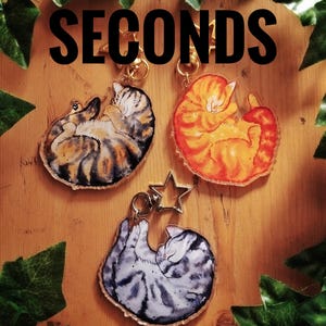 May include: Three acrylic keychains featuring illustrations of curled-up sleeping cats in brown tabby, orange tabby, and grey tabby patterns. Each keychain includes a gold-toned star-shaped clasp. The word SECONDS is printed in large black letters.