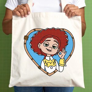 K&ouml;nnte beinhalten: Eine cremefarbene Tragetasche mit einer Cartoon-Illustration einer Figur mit roten Haaren, gr&uuml;nen Augen und einem wei&szlig;en und gelben Outfit. Die Figur befindet sich in einem herzf&ouml;rmigen Rahmen mit blauem Hintergrund.