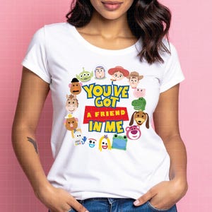 May include: A white short-sleeved t-shirt featuring a circular arrangement of various cartoon characters surrounding the text "YOU'VE GOT A FRIEND IN ME" printed in blue and yellow letters over a red rectangular block on the center of the chest.