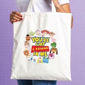 May include: A white canvas tote bag featuring a circular arrangement of colorful cartoon character faces surrounding the text "YOU'VE GOT A FRIEND IN ME" printed in bold blue and red lettering on the center of the fabric surface.