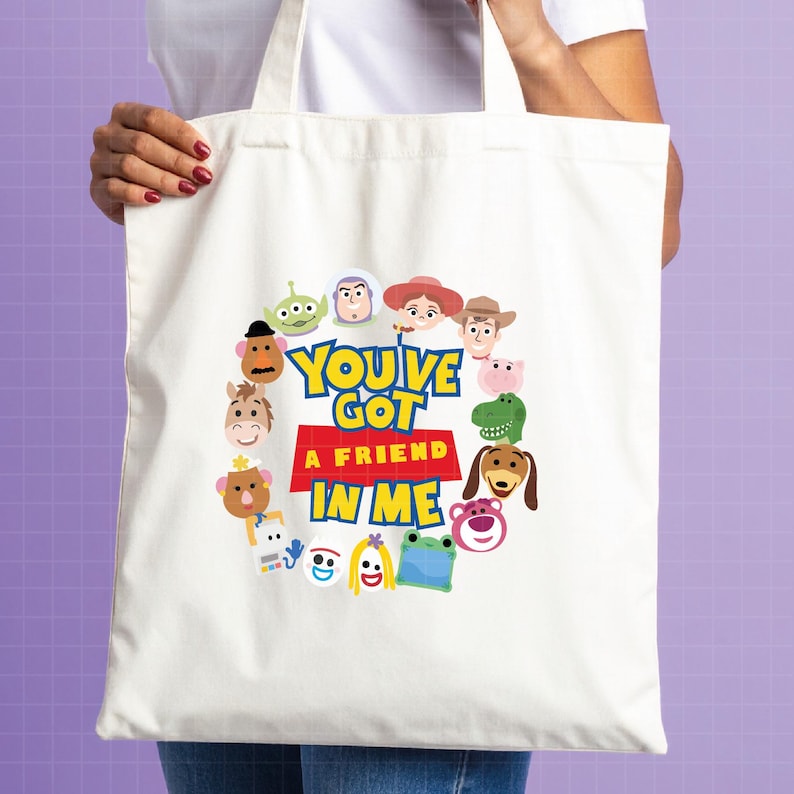 May include: A white canvas tote bag featuring a circular arrangement of colorful cartoon character faces surrounding the text "YOU'VE GOT A FRIEND IN ME" printed in bold blue and red lettering on the center of the fabric surface.
