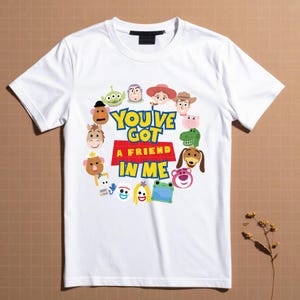 May include: A white short-sleeve t-shirt featuring a circular arrangement of various cartoon character faces surrounding the text "YOU'VE GOT A FRIEND IN ME" printed in blue and yellow letters on a red background, with a black neck label.