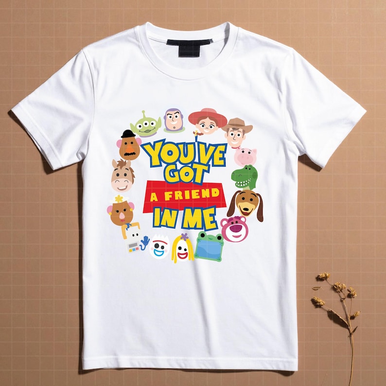 May include: A white short-sleeve t-shirt featuring a circular arrangement of various cartoon character faces surrounding the text "YOU'VE GOT A FRIEND IN ME" printed in blue and yellow letters on a red background, with a black neck label.