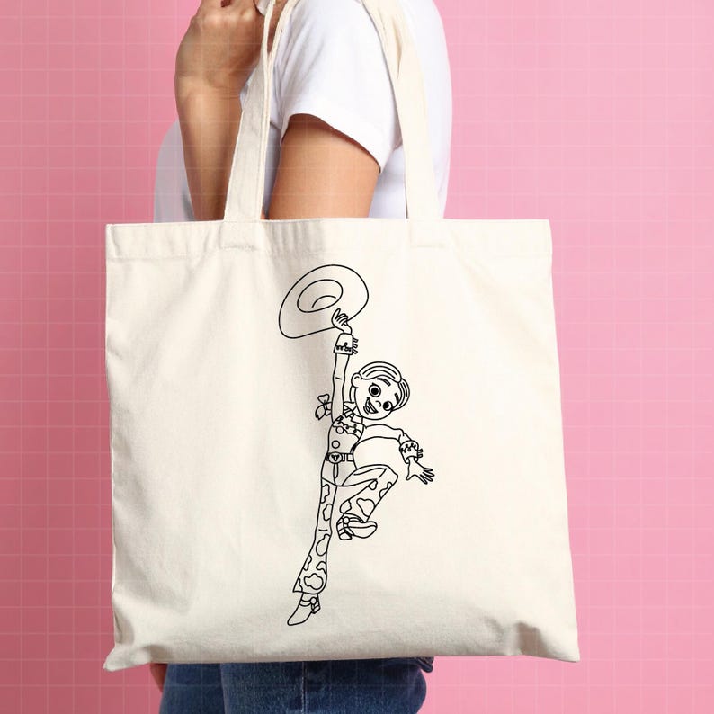 May include: A cream-colored tote bag featuring a black outline illustration of a cartoon cowgirl holding a hat. The cowgirl is wearing a cow-print outfit and is in a jumping pose. The bag has two cream-colored handles.