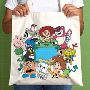 Puede incluir: Bolso tote color crema con un gr&aacute;fico colorido de personajes de Toy Story, incluyendo a Woody, Buzz Lightyear y Jessie. El dise&ntilde;o presenta varios personajes de la pel&iacute;cula animada. El bolso es sostenido por una persona.