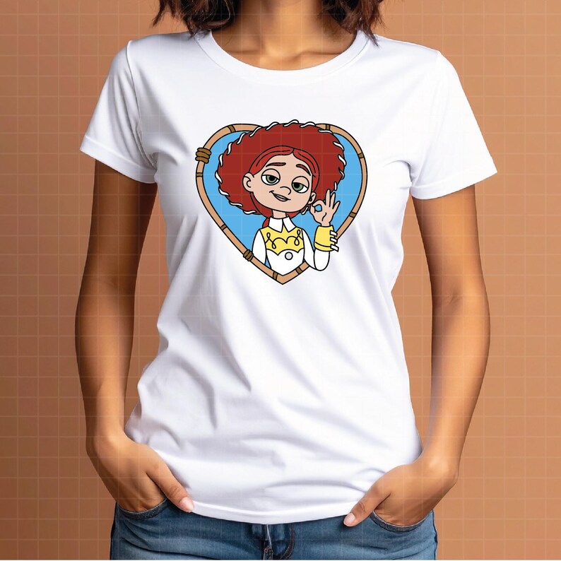 K&ouml;nnte beinhalten: Wei&szlig;es T-Shirt mit einer Cartoon-Illustration einer Figur mit roten Haaren, die in einem herzf&ouml;rmigen Rahmen dargestellt ist. Die Figur tr&auml;gt ein gelb-wei&szlig;es Outfit und macht eine &bdquo;Okay&ldquo;-Handgeste. Das Design ist mittig auf dem T-Shirt platziert.