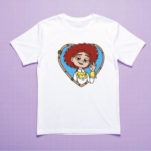 K&ouml;nnte beinhalten: Wei&szlig;es T-Shirt mit einer Cartoon-Illustration eines Charakters mit roten Haaren, innerhalb eines herzf&ouml;rmigen Rahmens. Die Figur macht die "Okay"-Handgeste. Das Design ist auf dem T-Shirt zentriert.