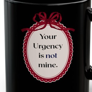 May include: Black ceramic mug with a white oval design featuring the text "Your Urgency is not mine." The oval is framed with a red scalloped border and a red bow at the top. The mug has a curved handle.