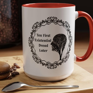 May include: White ceramic mug with a red interior and handle. The mug features a black decorative frame with the text "Tea First Existential Dread Later" and a tree-shaped silhouette of a face.