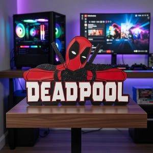 May include: A red and black Deadpool figure with the word "DEADPOOL" in white block letters. The figure is posed with crossed swords and is displayed on a wooden table. A computer and monitor are in the background.