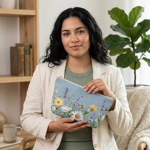 Puede incluir: Un neceser azul claro con un diseño floral con margaritas amarillas y blancas, y lavanda morada. El neceser tiene una cremallera dorada. Una persona sostiene el neceser.