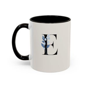 May include: White ceramic mug with a black handle and interior. The letter "E" is displayed in black with a blue floral design. The mug is a standard size for tea or coffee.