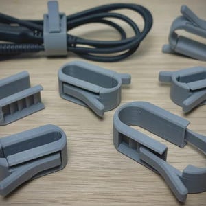 May include: Gray plastic cable clips and a cable are arranged on a light wood surface. The clips are in various sizes and shapes, designed to organize and secure cords. One clip is holding the cable.