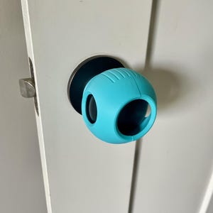 May include: A light blue door knob cover, spherical in shape, installed on a white door. The cover has a ridged texture and two large openings. The door hardware is silver.