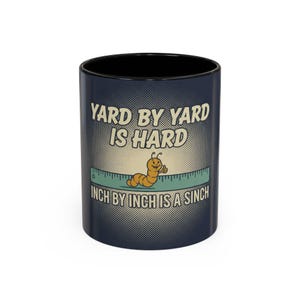 May include: A navy blue ceramic mug with a black interior. The mug features the text "YARD BY YARD IS HARD, INCH BY INCH IS A SINCH" with a cartoon worm giving a thumbs up on a ruler.