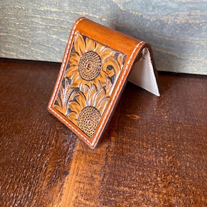 May include: A brown leather money clip with a sunflower design. The clip features two sunflowers and leaves, with white stitching around the edges. The interior is a light beige color.