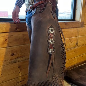 May include: Brown leather chaps with decorative silver and red accents. The chaps have a fringed bottom and are worn with a decorative belt. The chaps are a western wear item.