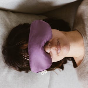 May include: A solid purple sleep mask with a smooth fabric finish. The mask features a small white rectangular tag on the lower edge with a black safety pin icon and the text MARU printed in black, positioned over the eyes.