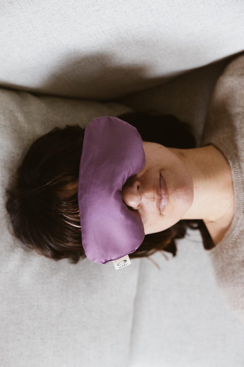 May include: A solid purple sleep mask with a smooth fabric finish. The mask features a small white rectangular tag on the lower edge with a black safety pin icon and the text MARU printed in black, positioned over the eyes.