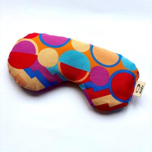 May include: A fabric sleep mask featuring a vibrant geometric pattern of circles and angular shapes in shades of red, orange, blue, and magenta. A small white fabric tag on the side displays a safety pin icon and the text MARV in black ink.