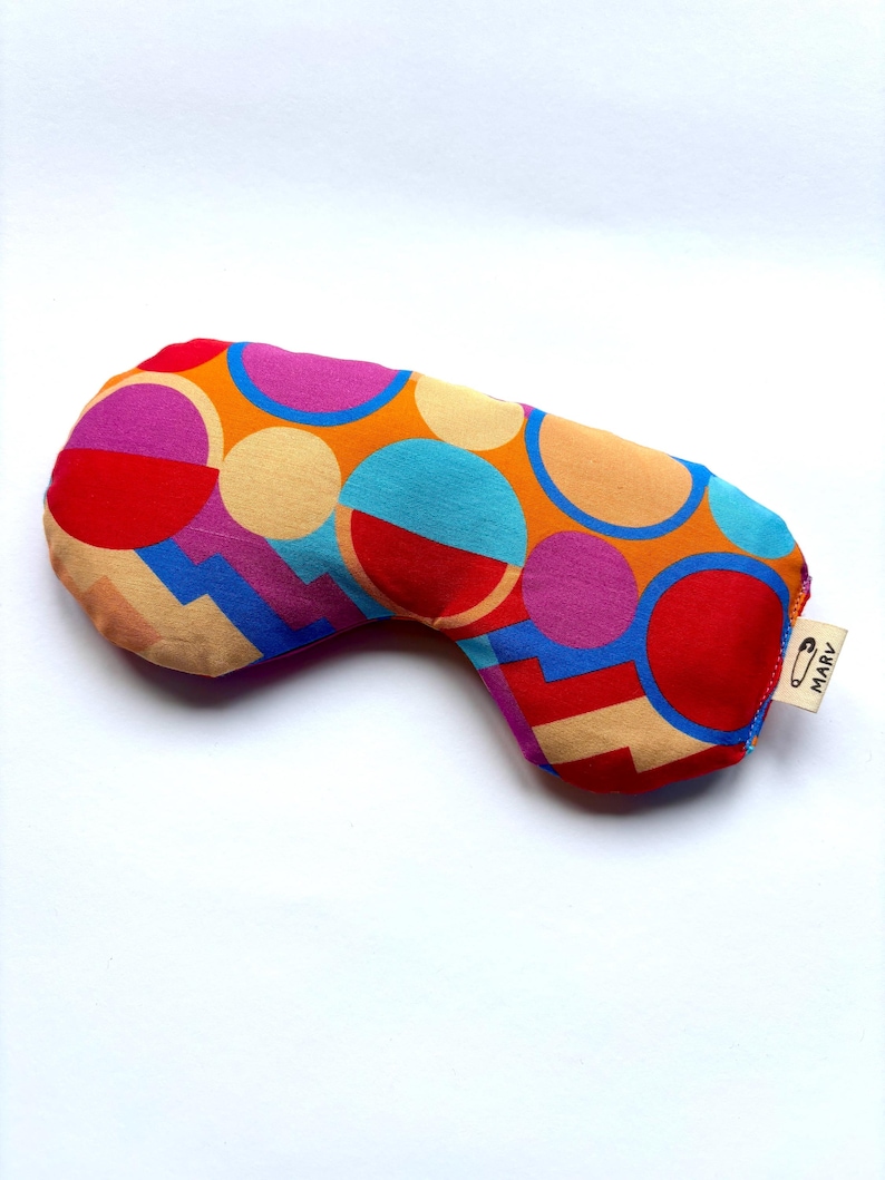 May include: A fabric sleep mask featuring a vibrant geometric pattern of circles and angular shapes in shades of red, orange, blue, and magenta. A small white fabric tag on the side displays a safety pin icon and the text MARV in black ink.